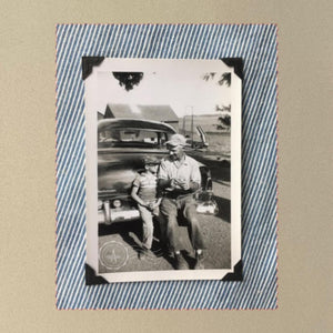A vintage photo of a boy and his son sitting on the bumper of an old car.