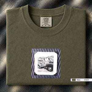 Olive colored folded t-shirt with vintage photo of mechanics and cars on front of shirt.
