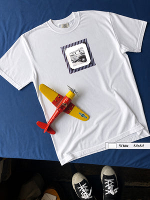 White t-shirt on table with vintage photo of mechanics and cars on front of shirt.